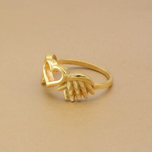 May include: A gold-colored ring featuring a heart-shaped design with a wing-like embellishment. The ring is set against a neutral background, highlighting its intricate details and polished finish. The ring's design is delicate and feminine.
