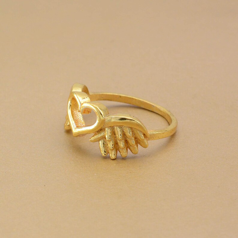 May include: A gold-colored ring featuring a heart-shaped design with a wing-like embellishment. The ring is set against a neutral background, highlighting its intricate details and polished finish. The ring's design is delicate and feminine.