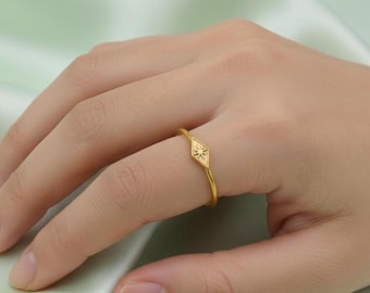 May include: A gold ring featuring a diamond-shaped centerpiece with a starburst design. The ring is worn on a finger, showcasing its delicate band and intricate detailing. The ring's warm gold color contrasts with the skin tone.