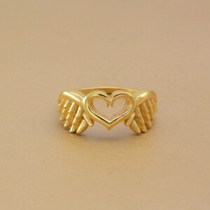 May include: A gold-colored ring featuring a heart-shaped center and wing-like designs on either side. The ring has a polished finish and is set against a neutral background. The ring is a piece of jewelry.