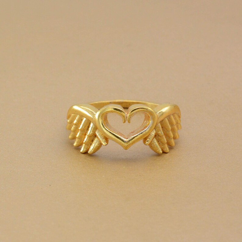 May include: A gold-colored ring featuring a heart-shaped center and wing-like designs on either side. The ring has a polished finish and is set against a neutral background. The ring is a piece of jewelry.