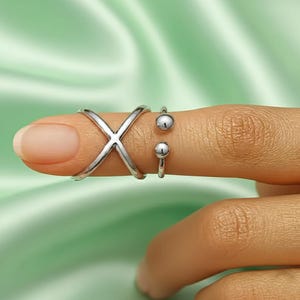 May include: A silver-colored ring with an X-shaped design is worn on a finger. Two additional silver rings with small ball accents are also worn on the same finger. The rings are set against a soft green background.