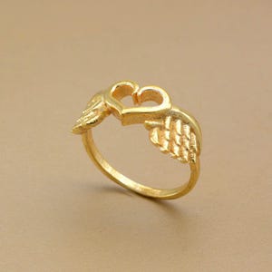 May include: A gold-colored ring featuring a heart-shaped design with two wings on either side. The ring is made of a shiny, metallic material and is set against a neutral background. The ring's design is detailed and intricate.