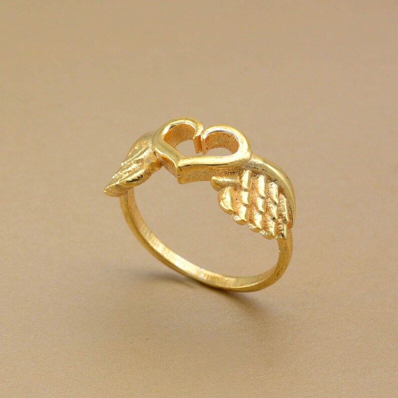 May include: A gold-colored ring featuring a heart-shaped design with two wings on either side. The ring is made of a shiny, metallic material and is set against a neutral background. The ring's design is detailed and intricate.