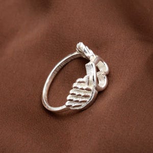 May include: A silver ring with a unique design. The ring features a stylized feather or leaf motif, with intricate detailing. The ring is set against a brown background, highlighting its metallic shine. The ring's design is both elegant and eye-catching.