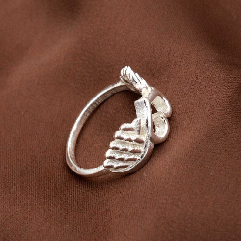 May include: A silver ring with a unique design. The ring features a stylized feather or leaf motif, with intricate detailing. The ring is set against a brown background, highlighting its metallic shine. The ring's design is both elegant and eye-catching.