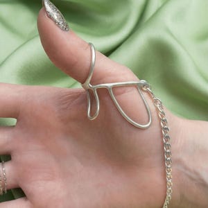 May include: A silver-colored hand chain ring. The ring is attached to a chain that wraps around the wrist. The ring is a unique, abstract design. The background is a light green fabric.