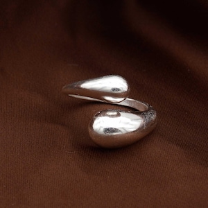 May include: A silver-colored ring with a unique design. The ring features two teardrop-shaped elements that curve around the finger. The ring is set against a brown background.