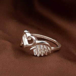 May include: A silver ring featuring a heart-shaped design. The ring has a delicate band and a detailed heart motif, with a textured wing-like element. The ring is displayed on a brown background.