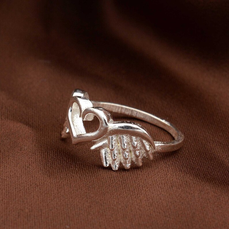 May include: A silver ring featuring a heart-shaped design. The ring has a delicate band and a detailed heart motif, with a textured wing-like element. The ring is displayed on a brown background.