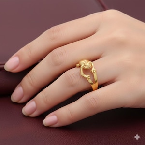 Könnte beinhalten: Ein goldfarbener Ring mit einem Frosch-Design. Der Frosch befindet sich oben auf dem Ring, mit seinen Armen, die sich zum Band erstrecken. Der Ring wird an einem Finger getragen und zeigt seine komplizierten Details und sein glänzendes Finish.