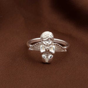 May include: A silver ring featuring a detailed angel figurine with folded hands and outstretched wings. The angel is kneeling, with a serene expression. The ring is set against a brown background.