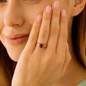 May include: A gold ring with a red gemstone. The ring is on a finger. The ring has a band with a textured design. The gemstone is oval-shaped and set in a gold setting. The ring is a piece of jewelry.
