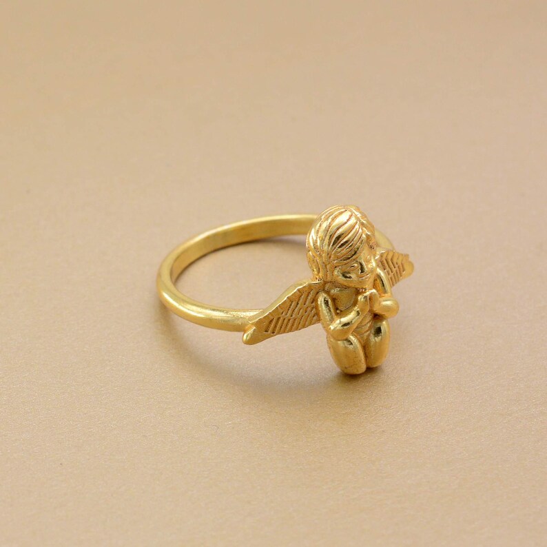 May include: A gold-colored ring featuring a small cherub with detailed wings and a serene expression. The ring's band is smooth and circular, and the cherub is the focal point of the design. The ring is set against a neutral background.