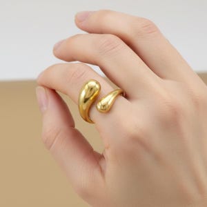 Puede incluir: Un anillo abierto de tono dorado que presenta dos extremos lisos y pulidos en forma de lágrima que se curvan uno hacia el otro. El metal tiene un acabado brillante y reflectante, con un diseño de formas fluidas, orgánicas y redondeadas.