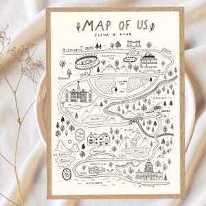 Op de afbeelding: Een ingelijste, gebroken witte print met een zwarte lijntekeningkaart getiteld MAP OF US met de namen ELENA & RYAN. De illustratie toont diverse bezienswaardigheden, wegen, bomen en wateren, met specifieke locaties aangeduid op de kaart.