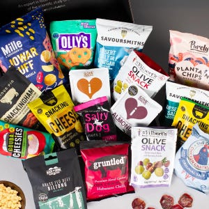 May include: A selection of snack bags and boxes, including cheese puffs, plantain chips, olive snacks, and beef biltong. The bags are colourful and feature various designs and logos.