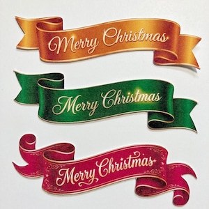 May include: Three decorative paper banners in gold, green, and red, each featuring the text Merry Christmas in elegant cursive script. The banners have a curled ribbon design with gold-toned edges and intricate scrollwork on the red banner.