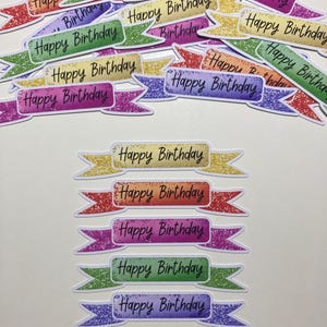 May include: A selection of colourful, glittery banner stickers with the words "Happy Birthday" in a handwritten style. The banners are in various colours, including gold, red, purple, green, and blue.
