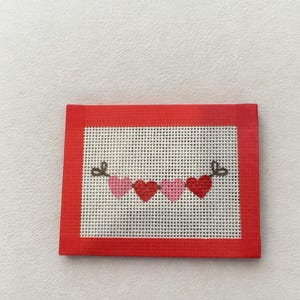 May include: A small, rectangular needlepoint featuring a garland of hearts. The hearts are in shades of pink and red, strung across a white mesh background. The border is a solid red, creating a framed effect. Two small bows are at each end of the garland.