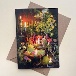 May include: A rectangular greeting card featuring a dark, lush botanical scene with lit candles, small red-capped mushrooms, green ferns, yellow flowers, and a string of pearls. The image is decorated with small, bright yellow star-shaped icons.