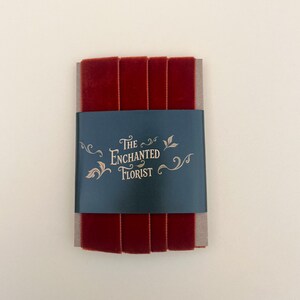 Op de afbeelding: Een bundel diep rood fluwelen lint, verpakt met een marineblauwe band met de tekst "The Enchanted Florist" in gouden letters. Het lint is netjes opgestapeld en toont zijn rijke kleur en textuur.