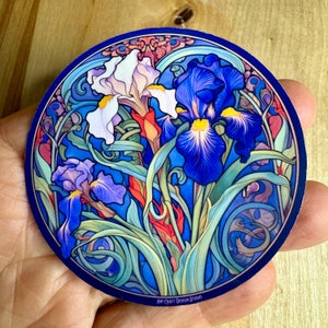 May include: A round sticker featuring a colorful art nouveau style illustration of three iris flowers with blue, white, and purple petals. The background is a deep blue with intricate swirling patterns. The sticker has the text "Art Craft Design Studio" at the bottom.