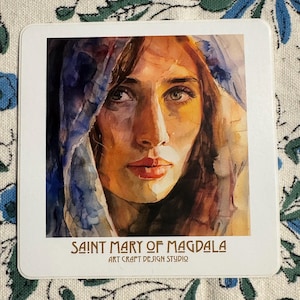 May include: Watercolor painting of a woman with long brown hair wearing a blue headscarf. The painting is titled "Saint Mary of Magdala" with the text "Art Craft Design Studio" below the title.
