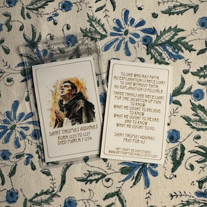 May include: A prayer card featuring a watercolor illustration of Saint Thomas Aquinas, a Catholic saint. The card includes a quote from Saint Thomas Aquinas and his birth and death dates. The card is printed on white cardstock with gold lettering.