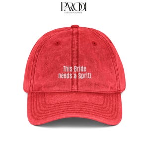 May include: Red baseball cap with the embroidered text "This Bride needs a Spritz." The cap has a curved brim and a fabric strap closure. The hat is a solid red color.