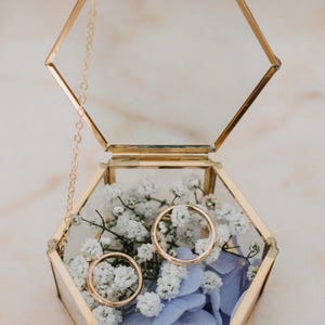Può includere: Una scatola di vetro esagonale con cornice dorata con due fedi nuziali d'oro all'interno. La scatola è aperta, rivelando fiori bianchi e petali blu. Una delicata catena d'oro è attaccata al coperchio.