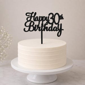 Może przedstawiać: Białe ciasto z kremem na białym stojaku na ciasto, zwieńczone czarnym topperem z napisem "Happy 30th Birthday". Ciasto ma poziome, teksturowane linie.