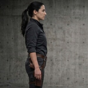 May include: A person wearing a dark gray button-down shirt, dark gray pants, and a brown leather holster with a gun. The person has dark hair pulled back in a ponytail and is standing in front of a gray concrete wall.