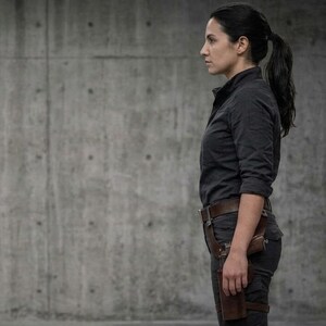 May include: A person in a dark gray button-up shirt and dark gray pants, with a brown leather holster and a gun on their right leg. The person is standing in front of a gray concrete wall.