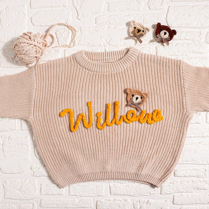 May include: A light beige knit sweater with the name "Willows" in yellow script. The sweater features a small teddy bear applique. Two additional bear appliques and a ball of yarn are also visible.