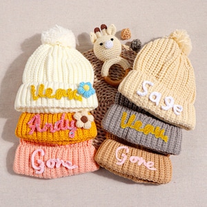 May include: Six knitted hats with pom-poms, each with a different name embroidered on them in colourful yarn. The names are: Andy, Gone, Gone, Sage, Lena, and Lena.