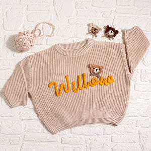 May include: A light beige knit sweater with the name "Willows" in yellow script. The sweater features a small teddy bear applique and is accompanied by a ball of yarn and additional bear appliques. The sweater is displayed on a white brick background.