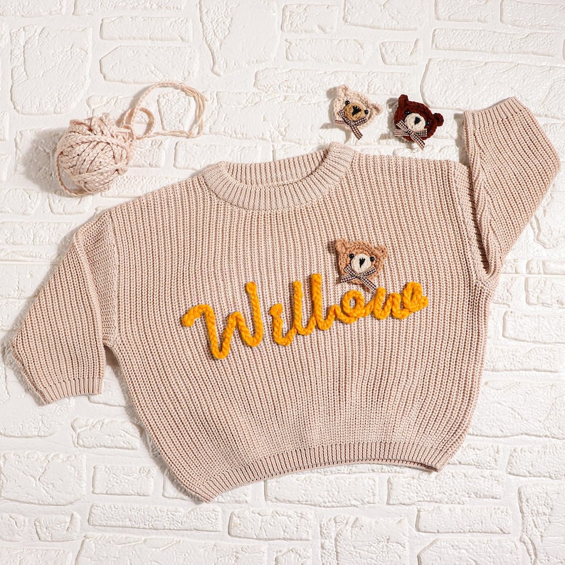 May include: A light beige knit sweater with the name "Willows" in yellow script. The sweater features a small teddy bear applique and is accompanied by a ball of yarn and additional bear appliques. The sweater is displayed on a white brick background.