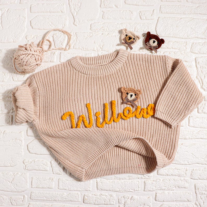 May include: A light beige baby sweater with the name "Willow" in yellow yarn. The sweater has a ribbed knit design and is decorated with three small teddy bear appliques. A ball of yarn and two additional teddy bear appliques are also present.