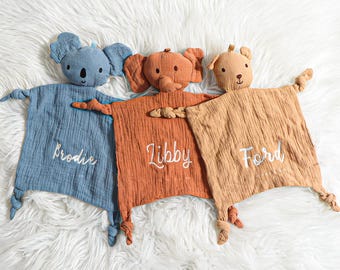 May include: Three plush animal security blankets with embroidered names. The blankets are blue, orange, and brown. The animals are a koala, an elephant, and a dog.