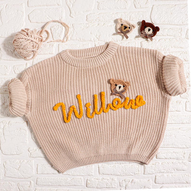 May include: A light beige knit sweater with the name "Willow" in yellow script. The sweater features a teddy bear applique and two additional bear appliques. A ball of yarn is in the upper left corner.