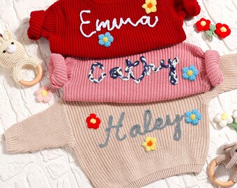 May include: Three knitted sweaters with embroidered names, Emma, Cathy, and Haley. Each sweater has a different color: red, pink, and beige. The sweaters are decorated with colorful flowers.