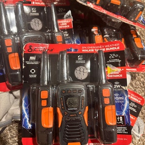 Peut inclure: Un lot de trois talkies-walkies Cobra avec des accents orange et noir. Les talkies-walkies sont étanches et ont une portée allant jusqu'à 64 kilomètres. L'emballage porte la mention "3PK Emergency Weather Walkie Talkie Bundle" et "22 Channels 121 Privacy Codes".