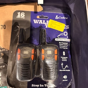 以下が含まれることがあります： オレンジ色のボタンとデジタルディスプレイが付いた、黒いコブラのトランシーバー2台。パッケージは青と白のデザインで、「トランシーバー」と「つながりましょう」の文字が書かれています。パッケージには、26kmの範囲、天気予報、充電式バッテリー、マイクロUSB充電など、製品の機能に関する情報も含まれています。