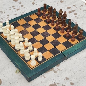 May include: A wooden chess set with a green border. The chessboard is folded open, revealing a checkerboard pattern of light and dark squares. Chess pieces are arranged on the board, with white and brown pieces facing each other.