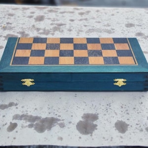 Puede incluir: Un juego de ajedrez de madera cerrado, de color verde azulado. El tablero es un patrón de cuadros marrones y negros. El conjunto tiene cierres y bisagras doradas. El tablero es de madera.