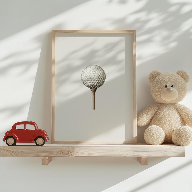 May include: A framed print featuring a golf ball on a tee. The print is displayed on a wooden shelf with a red toy car and a beige teddy bear. The background is a white wall with shadows.