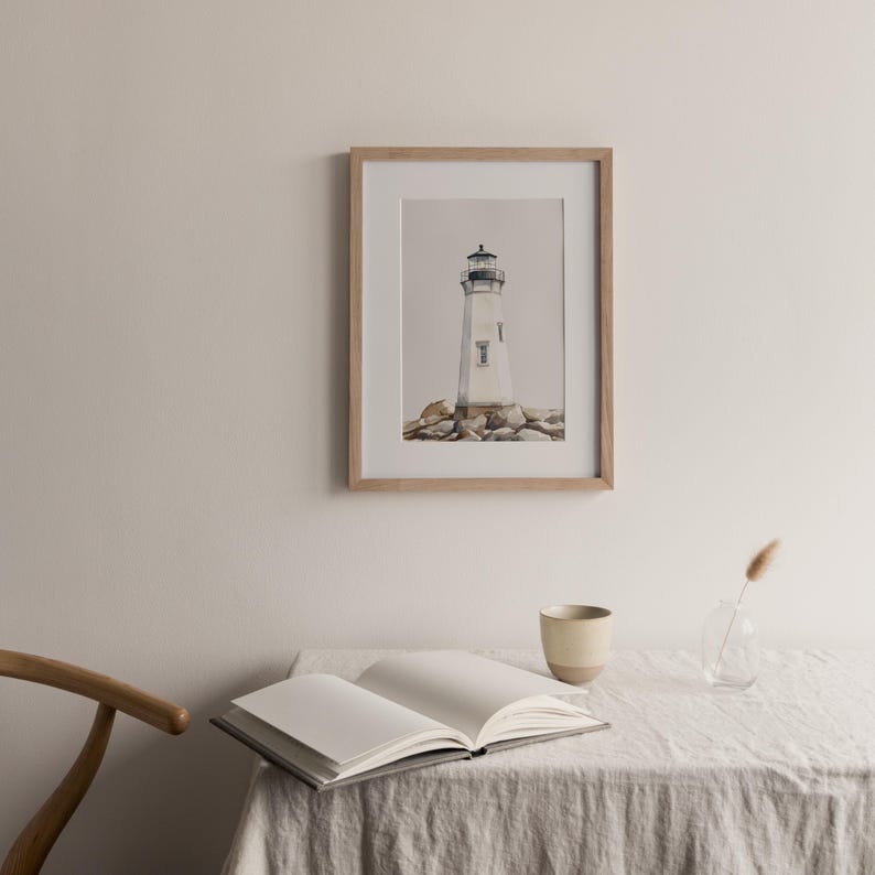 May include: A framed print of a lighthouse, set against a neutral background. The lighthouse is white with a black top, resting on a rocky base. The frame is light wood. A book, cup, and vase are on a table.