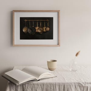 Puede incluir: Una l&aacute;mina enmarcada que muestra un estante met&aacute;lico horizontal con siete sartenes de cobre y metal oscuro colgadas sobre un fondo negro. El marco es de madera clara y la l&aacute;mina se exhibe sobre un libro abierto y una taza de cer&aacute;mica.