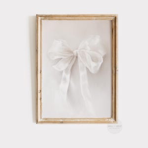 Pode incluir: Um laço de cetim branco amarrado em um nó, exibido em uma moldura de madeira clara. O laço está centralizado na moldura e o fundo é de cor creme claro.